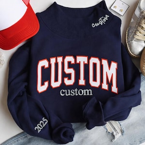 Può includere: Felpa blu navy con la scritta "CUSTOM" in grandi lettere bianche bordate di rosso. La parola "custom" è ricamata sotto. L'anno "2025" è ricamato sulla manica. Un cappellino da camionista rosso e bianco è visibile sullo sfondo.