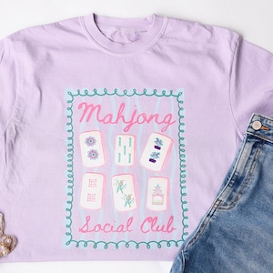 Puede incluir: Una camiseta de color lila claro con un gráfico de fichas de Mahjong y el texto "Mahjong Social Club" en rosa. El diseño está enmarcado por un borde turquesa. La camiseta se combina con vaqueros azules y joyas doradas.
