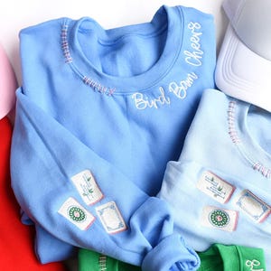 Puede incluir: Sudadera azul claro con detalles bordados y el texto "Bird Bam Cheers". La sudadera presenta parches bordados en la manga y costuras decorativas alrededor del cuello. Tambi&eacute;n se ven otras sudaderas de colores y una gorra blanca.