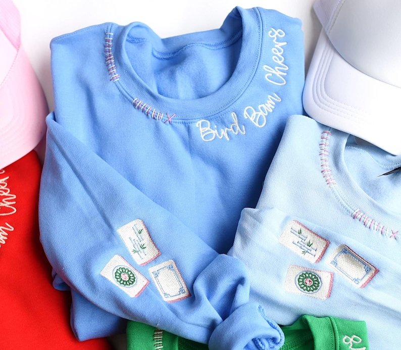 Puede incluir: Sudadera azul claro con detalles bordados y el texto "Bird Bam Cheers". La sudadera presenta parches bordados en la manga y costuras decorativas alrededor del cuello. Tambi&eacute;n se ven otras sudaderas de colores y una gorra blanca.