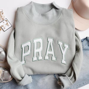 Könnte beinhalten: Hellgrünes Sweatshirt mit dem Wort "PRAY" in weißen Blockbuchstaben mit grünem Umriss. Das Sweatshirt hat einen Rollkragen und lange Ärmel. Ein hellrosa Hut und Jeans sind im Hintergrund.