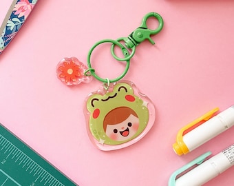 May include: A green frog keychain with a smiling face and a flower charm. The keychain is made of clear plastic and has a metal ring.