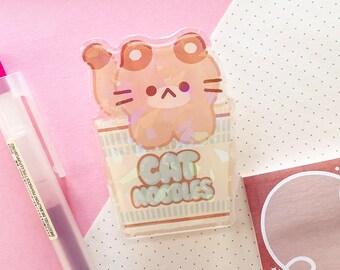 May include: A clear sticker with a cartoon cat inside a ramen noodle cup. The cat is brown and has a light pink nose. The cup says "Cat Noodles" in a light blue font.