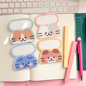 May include: Four cat-shaped sticky notes in pastel shades, each with a blank speech bubble. The cats are in orange and white, pink and grey, blue, and brown. There are also yellow and pink highlighters and a pink pen in the image.