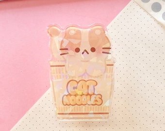 May include: A clear, iridescent sticker featuring a cartoon cat inside a container of noodles. The sticker has the words "CAT NOODLES" in a playful font. The cat is light brown and white.