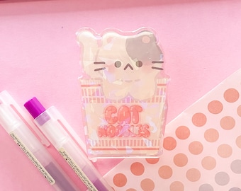 May include: A clear plastic sticker with a cartoon cat in a ramen noodle container. The cat is white with black spots and has a pink nose. The text "CAT NOODLES" is printed on the container.