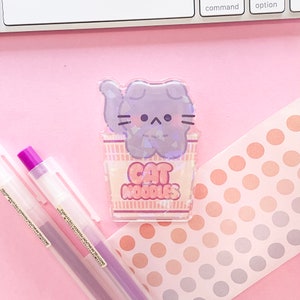 May include: A clear holographic sticker featuring a cartoon cat in a purple ramen bowl with the text "CAT NOODLES".