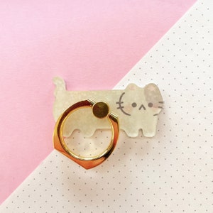 May include: A gold phone ring holder with a white cat design. The cat has a black nose and eyes and a white patch on its forehead.