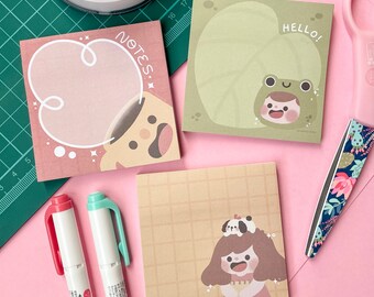May include: A set of three square sticky note pads with cute illustrations. The first pad has a pink background with a white cloud and a brown dog with a surprised expression. The second pad has a green background with a frog sitting on a lily pad. The third pad has a tan background with a grid pattern and a girl with a brown pigtail wearing a brown dress and a white hat with a brown pig on top. The text "NOTES:" is on the first pad and "HELLO!" is on the second pad.