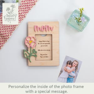 May include: Wooden photo frame with a pink flower cutout and the word "mom" on the top. The frame has a space for a photo and a personalized message inside. The message reads "Happy Mother's Day. We love and appreciate you so much. With all our love always. May and Dan, Maya and Olivia."