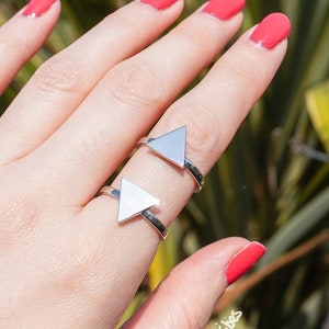 Op de afbeelding: Twee zilveren driehoekringen op een hand met rode nagellak. De ringen zijn open en verstelbaar. De hand is tegen een groene, bladerrijke achtergrond.