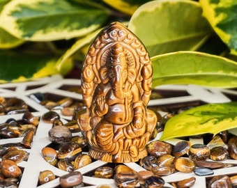 May include: A small, brown, carved Ganesha statue sits on a white wooden mandala with tiger's eye stones. The mandala is surrounded by green leaves.