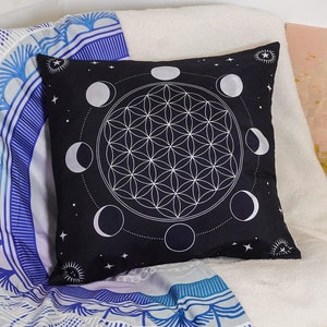 May include: Black cushion with a white flower of life design and a circle of moons around it. The cushion is on a white sofa with a blue and white tapestry.