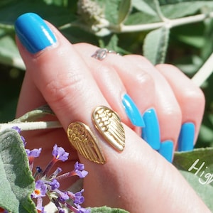 Op de afbeelding: Een gouden ring met een vleugelontwerp op de duim van een hand met blauwe nagellak. De hand houdt een tak van een plant met paarse bloemen vast.