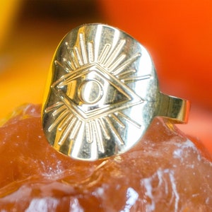 Puede incluir: Un anillo dorado con un diseño de ojo y rayos de sol. El anillo está sobre una superficie ámbar texturizada. Las palabras "High Goddess Vibes" son visibles en la imagen.