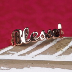 Könnte beinhalten: Roségoldener Ring mit dem Wort "Blessed" in kursiv.