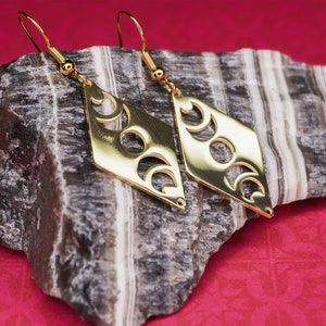 Può includere: Orecchini dorati a forma di diamante con un design a tripla luna tagliato. Gli orecchini sono esposti su una pietra grigia e bianca.