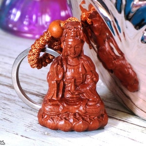 May include: A wooden keychain featuring a small, intricately carved statue of a seated figure. The figure is brown and has a serene expression. The keychain is attached to a silver ring and a brown cord.