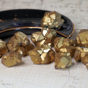 Può includere: Un gruppo di aggregati di pirite dorata, nota anche come oro degli stolti, disposti su una mappa vintage. Un piatto nero a forma di mezzaluna con bordi dorati e dettagli a stella contiene altra pirite. Il testo "High Goddess Vibes" è visibile.