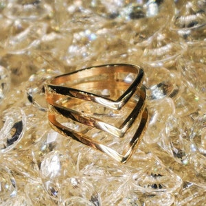 May include: A gold-toned ring with a triple chevron design. The ring is set against a background of clear, faceted crystals.