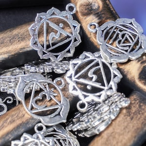 Może przedstawiać: Zbliżenie srebrnych metalowych zawieszek z misternymi wzorami. Zawieszki przedstawiają symbole i wzory związane z siedmioma czakrami. Zawieszki są ułożone na drewnianej powierzchni.