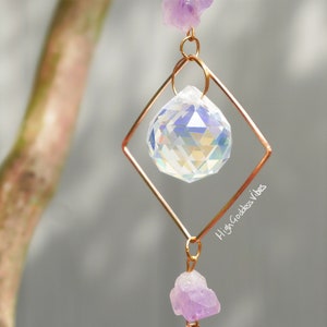 May include: A crystal suncatcher with a clear crystal prism hanging from a gold wire frame. The frame is shaped like a diamond and has amethyst crystals hanging from the bottom. The text "High Goddess Vibes" is printed on the wire frame.