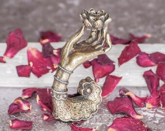 May include: A gold-toned metal hand sculpture holding a lotus flower. The sculpture is surrounded by red rose petals on a white surface.