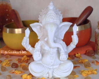May include: White Ganesha statue with a crown and a trunk, sitting in a meditative pose. The statue is surrounded by yellow flowers and crystals.