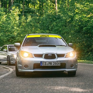 Op de afbeelding: Een zilveren Subaru Impreza WRX STI met een gele banner op de voorruit met de tekst "AKINA" en een kentekenplaat met de tekst "ES MK 2606" die over een kronkelende weg door een bos rijdt.