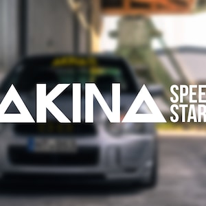 Può includere: Un'auto argentata con un triangolo bianco e la parola "AKINA" sul davanti. L'auto è parcheggiata davanti a un edificio con le parole "SPEED STARS" sul lato.