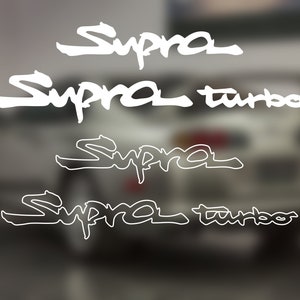 May include: Four white vinyl decals with the word "Supra" written in a stylized font. The top two decals also include the word "turbo" written in the same font.