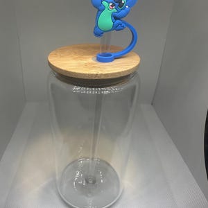 May include: A clear glass tumbler with a bamboo lid and a blue and pink Stitch character straw topper. The tumbler has a clear straw and is designed for cold beverages.