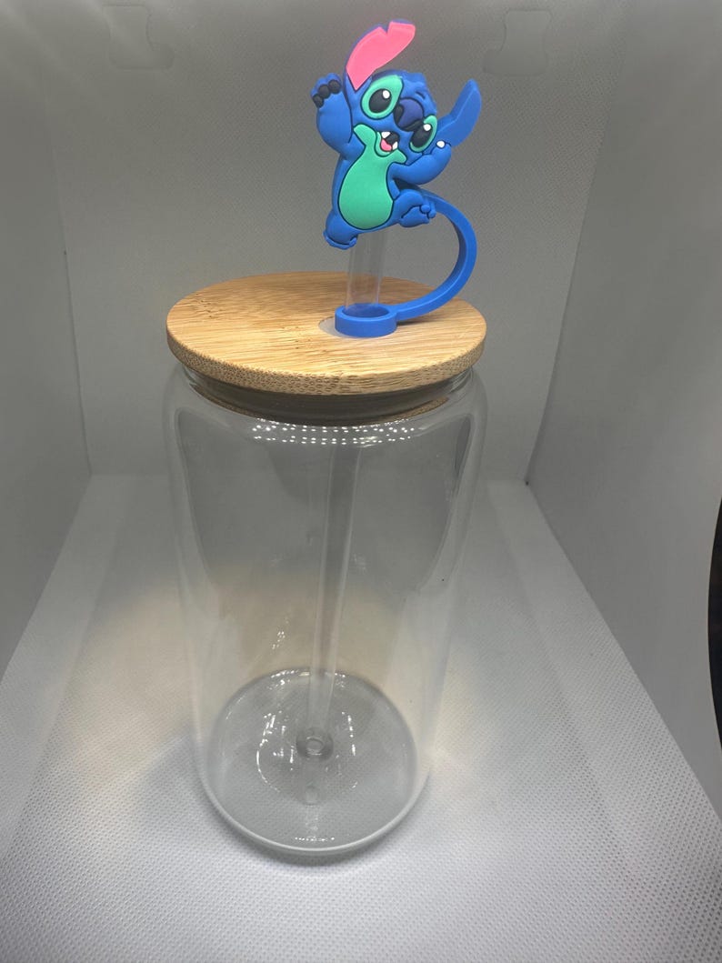 May include: A clear glass tumbler with a bamboo lid and a blue and pink Stitch character straw topper. The tumbler has a clear straw and is designed for cold beverages.