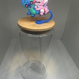 May include: A clear glass jar with a bamboo lid and a blue and green cartoon character topper. The topper features two characters embracing. The jar has a clear straw. The background is a light gray.