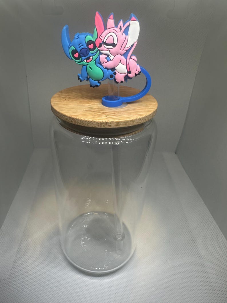 May include: A clear glass jar with a bamboo lid and a blue and green cartoon character topper. The topper features two characters embracing. The jar has a clear straw. The background is a light gray.