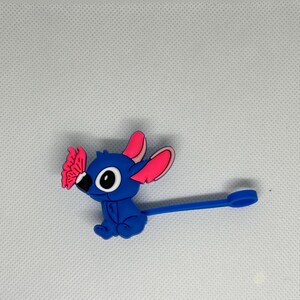 May include: A blue cartoon character with large eyes, pink-lined ears, and a butterfly on its nose. The character has a long, blue, stem-like appendage with a circular end. The figure is set against a white background.