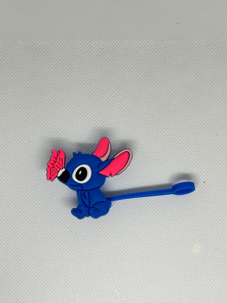 May include: A blue cartoon character with large eyes, pink-lined ears, and a butterfly on its nose. The character has a long, blue, stem-like appendage with a circular end. The figure is set against a white background.
