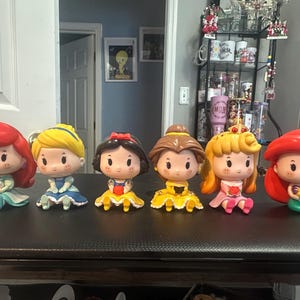 May include: Six cartoon figurines of Disney princesses, each with distinctive features like red, yellow, and brown hair, and colorful dresses. The figurines are arranged on a dark surface, showcasing their unique designs.