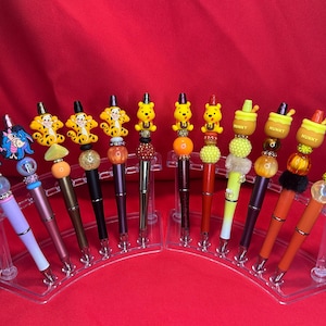 Puede incluir: Una colección de bolígrafos decorativos con adornos temáticos de personajes. Los bolígrafos tienen varios diseños, incluyendo Winnie the Pooh, Tigger y Eeyore. Los bolígrafos se exhiben en un soporte curvo transparente sobre un fondo rojo.
