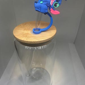 May include: A clear glass tumbler with a bamboo lid and a blue silicone straw holder featuring a cartoon character. The character is blue with pink accents and a long pink tongue. The tumbler has a clear straw.