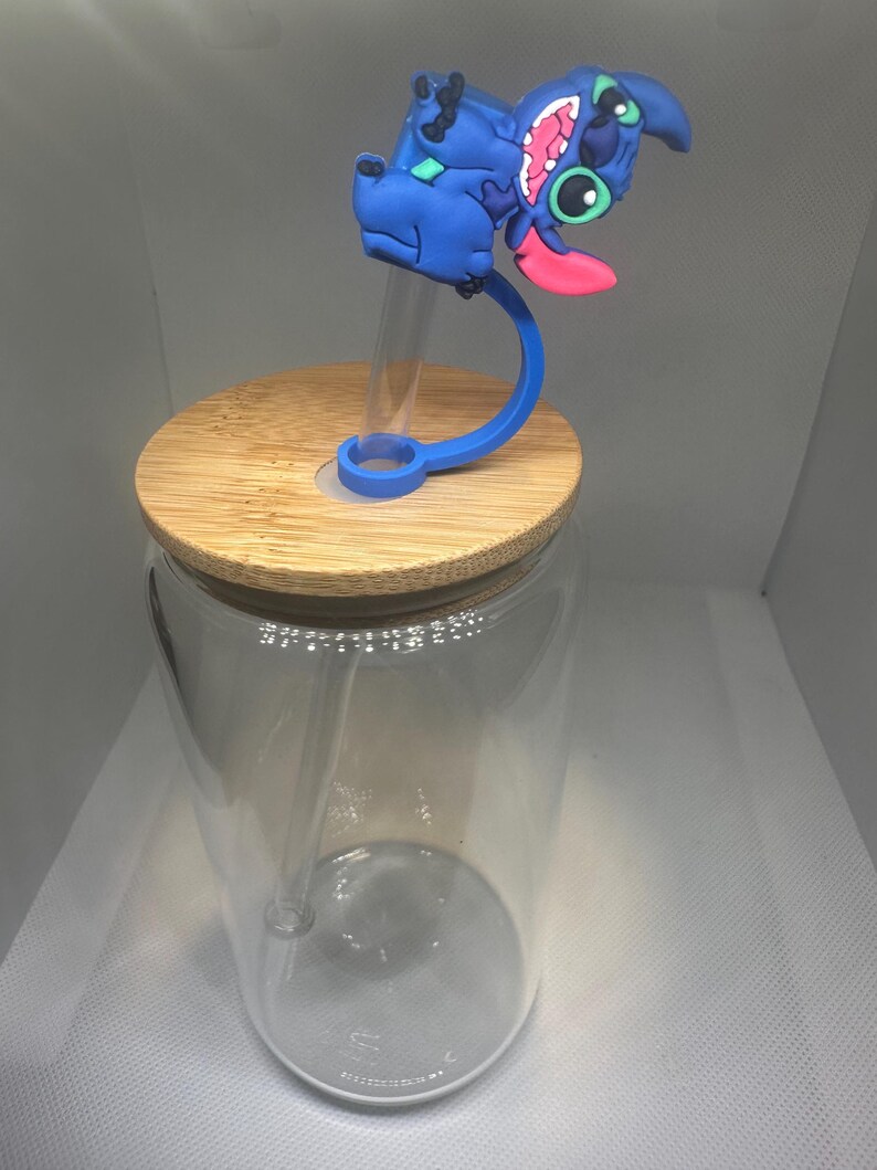 May include: A clear glass tumbler with a bamboo lid and a blue silicone straw holder featuring a cartoon character. The character is blue with pink accents and a long pink tongue. The tumbler has a clear straw.