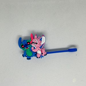 May include: A blue and green cartoon character embracing a pink cartoon character. The characters are attached to a blue plastic stick with a flat end. The characters have large eyes and are smiling. The background is white.