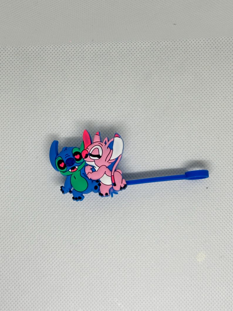 May include: A blue and green cartoon character embracing a pink cartoon character. The characters are attached to a blue plastic stick with a flat end. The characters have large eyes and are smiling. The background is white.