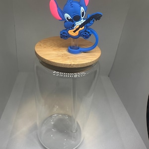 May include: A clear glass tumbler with a bamboo lid and a blue Stitch character topper playing a guitar. The topper has pink accents on the ears. The tumbler includes a clear straw.