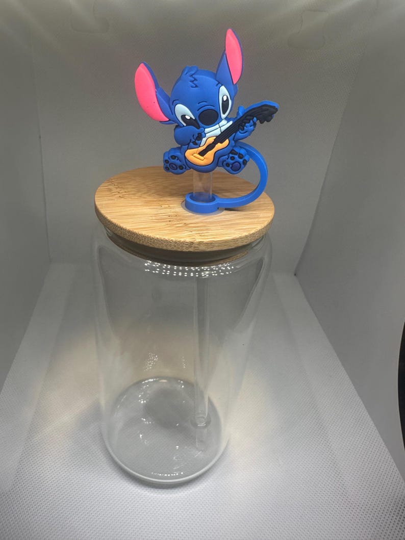 May include: A clear glass tumbler with a bamboo lid and a blue Stitch character topper playing a guitar. The topper has pink accents on the ears. The tumbler includes a clear straw.