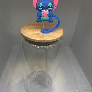 May include: A clear glass tumbler with a bamboo lid and a blue and pink Stitch character straw topper. The tumbler has a clear straw inside. The Stitch character is blue with pink ears and a green tongue.