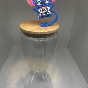 May include: A clear glass tumbler with a bamboo lid and a blue silicone straw topper featuring a cartoon character holding a sign that reads "FREE HUGS". The topper has pink ears and a blue body.