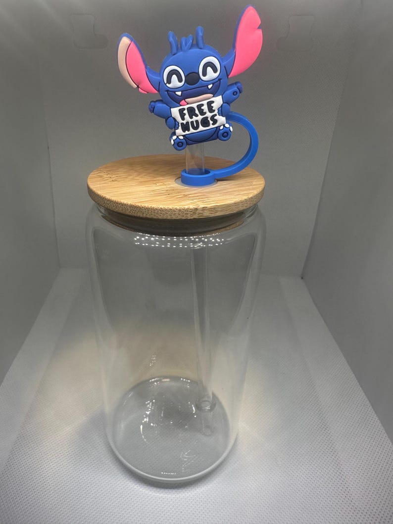 May include: A clear glass tumbler with a bamboo lid and a blue silicone straw topper featuring a cartoon character holding a sign that reads "FREE HUGS". The topper has pink ears and a blue body.