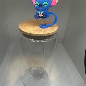 May include: A clear glass tumbler with a bamboo lid. A blue character figure with pink accents sits atop the lid. The character is holding a pink drink with a blue straw. The tumbler is empty.