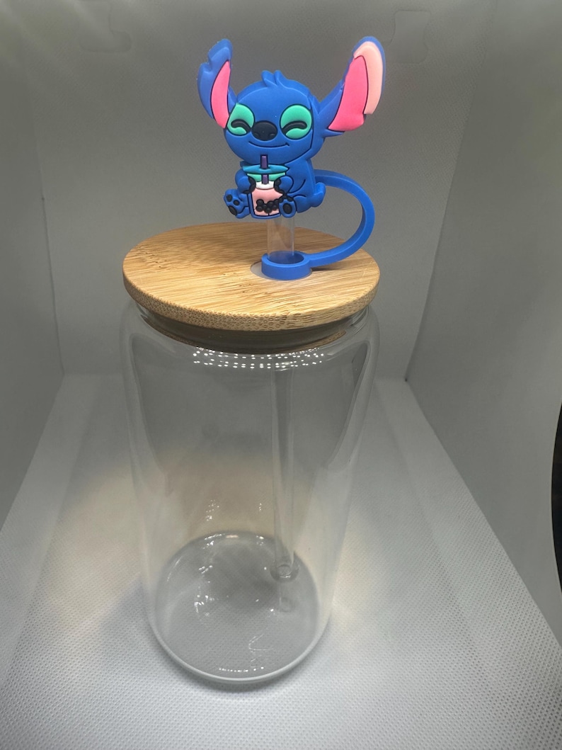 May include: A clear glass tumbler with a bamboo lid. A blue character figure with pink accents sits atop the lid. The character is holding a pink drink with a blue straw. The tumbler is empty.